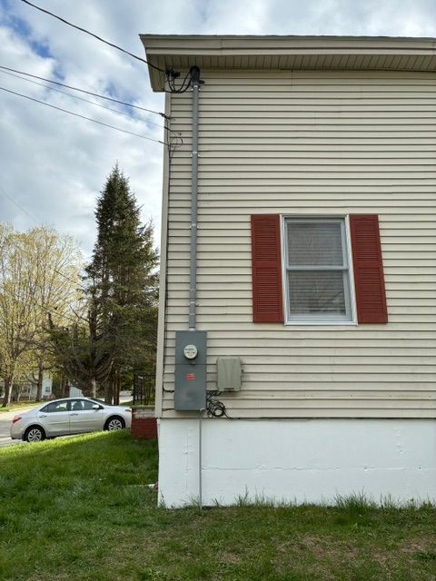 House side with beige siding, red shutters, utility meters, and grassy yard under a cloudy sky