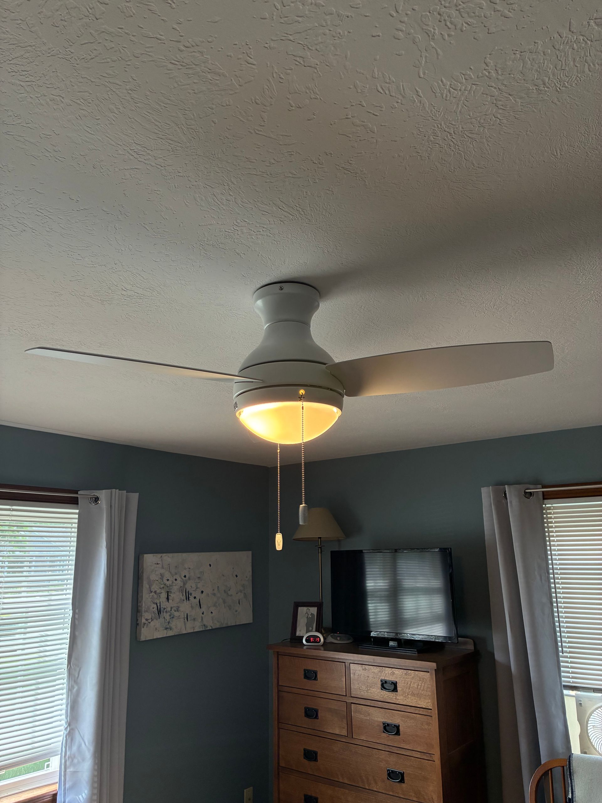 Bedroom ceiling fan with light on above a dresser, TV, and windows with blinds and curtains.