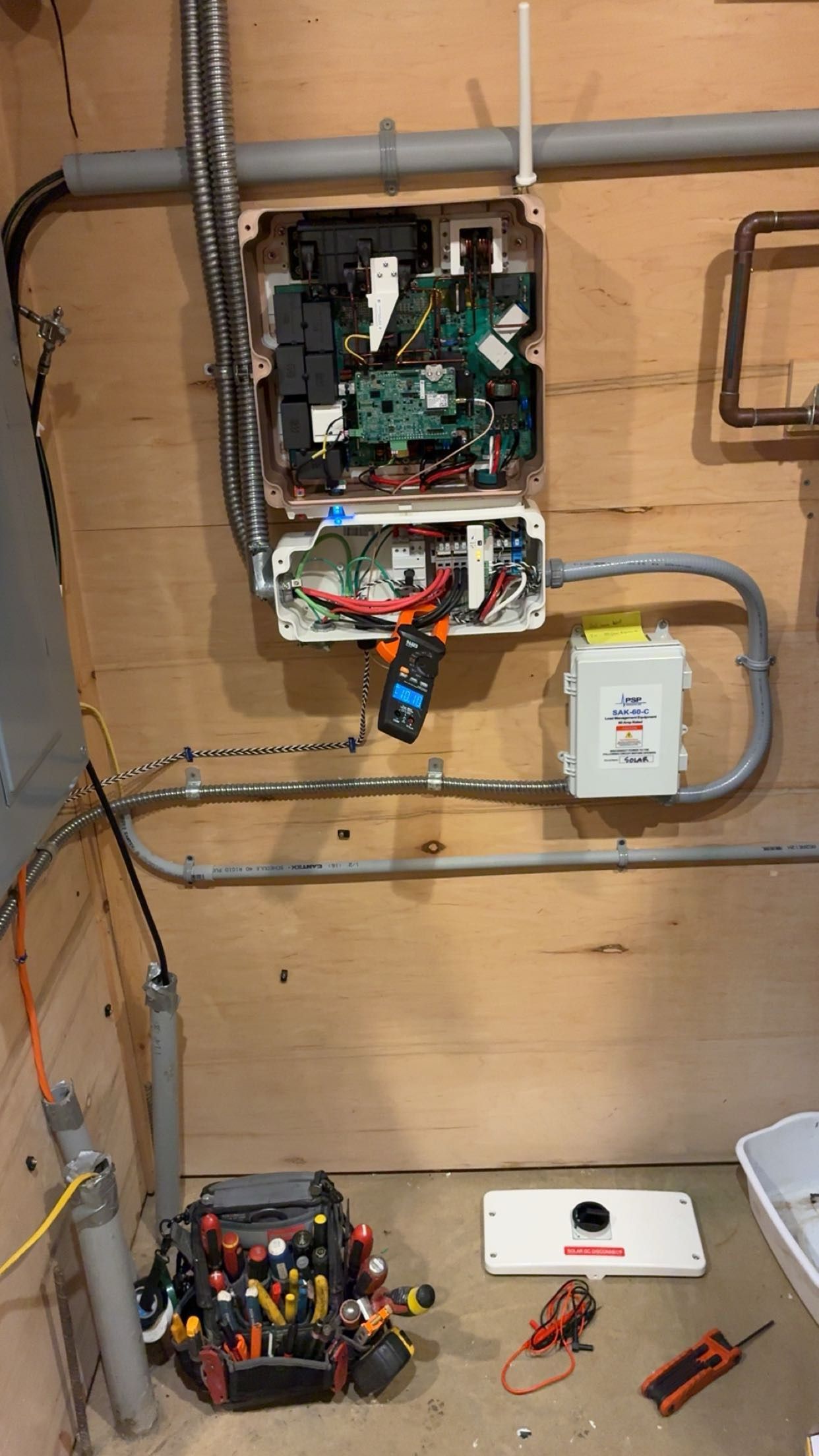 Wall-mounted electrical panel with exposed wiring and a small device below, in a utility room.