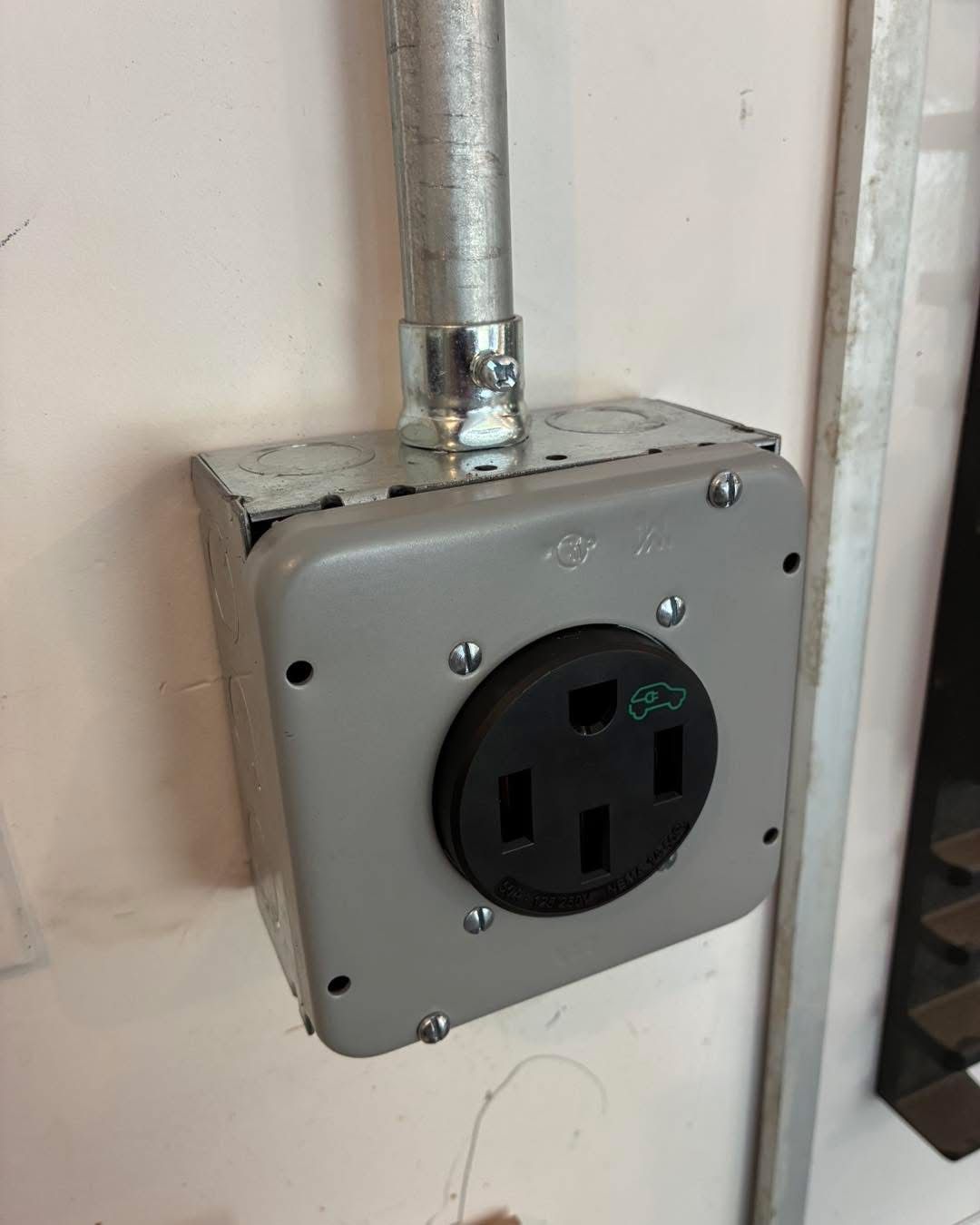 Gray electrical outlet box with a black socket mounted on a wall, connected to a metal conduit.