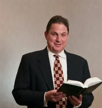 Man in a dark suit holding an open book against a plain background