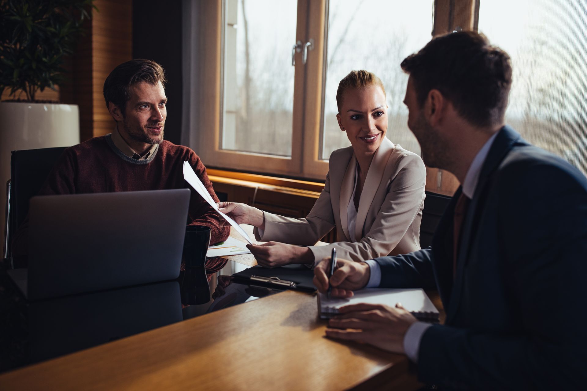 Three people in a meeting around a laptop at a wooden table near a window.