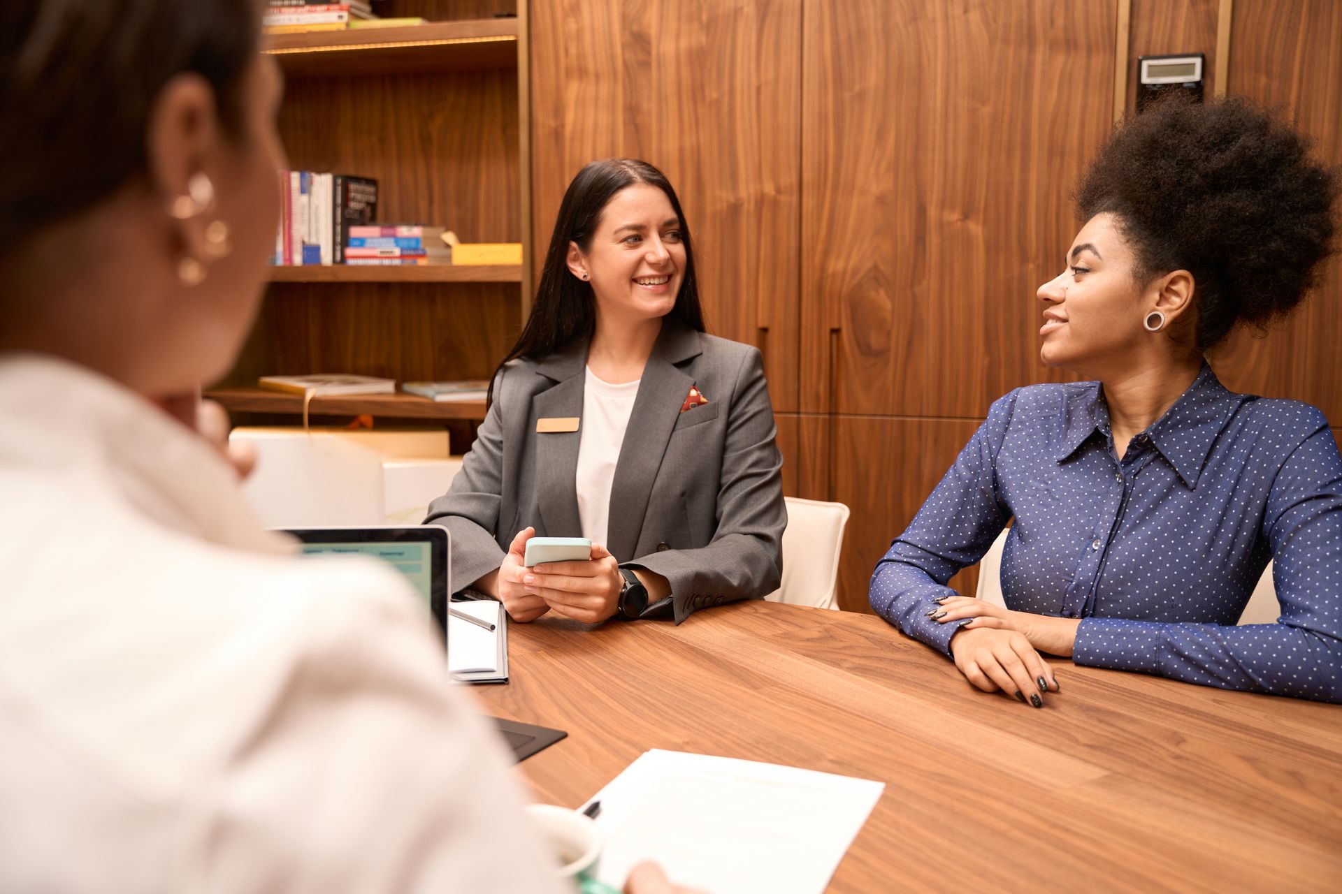 Three people in a meeting around a wooden table, smiling and holding documents.