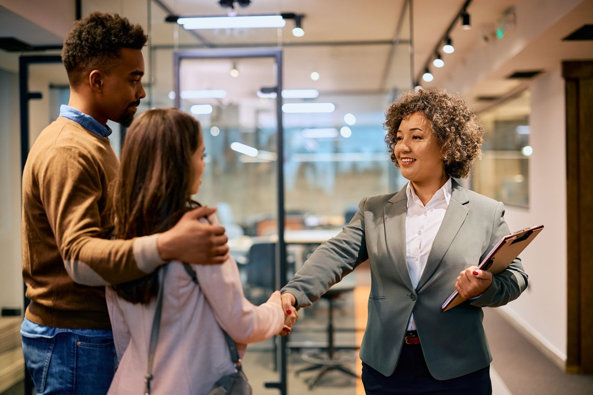 Two colleagues greet a businesswoman in an office hallway, smiling and shaking hands.