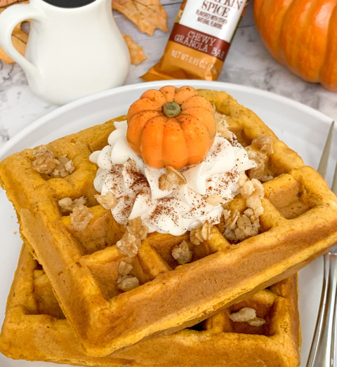 pumpkin spice waffles made with granola and topped with whipped cream, nuts and a candy pumpkin on a white plate