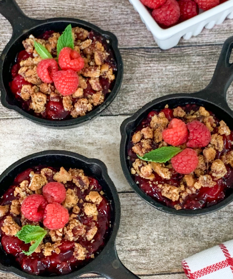 granola crumble in mini cast iron skilled topped with raspberries by a bowl of fresh raspberries in white container