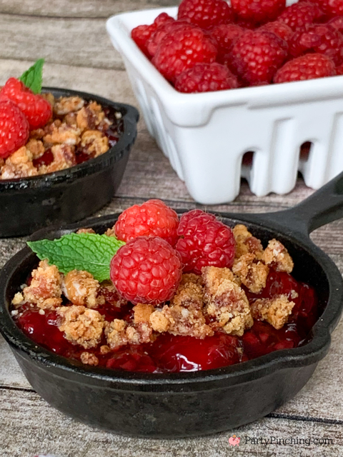 granola crumble in mini cast iron skilled topped with raspberries by a bowl of fresh raspberries in white container