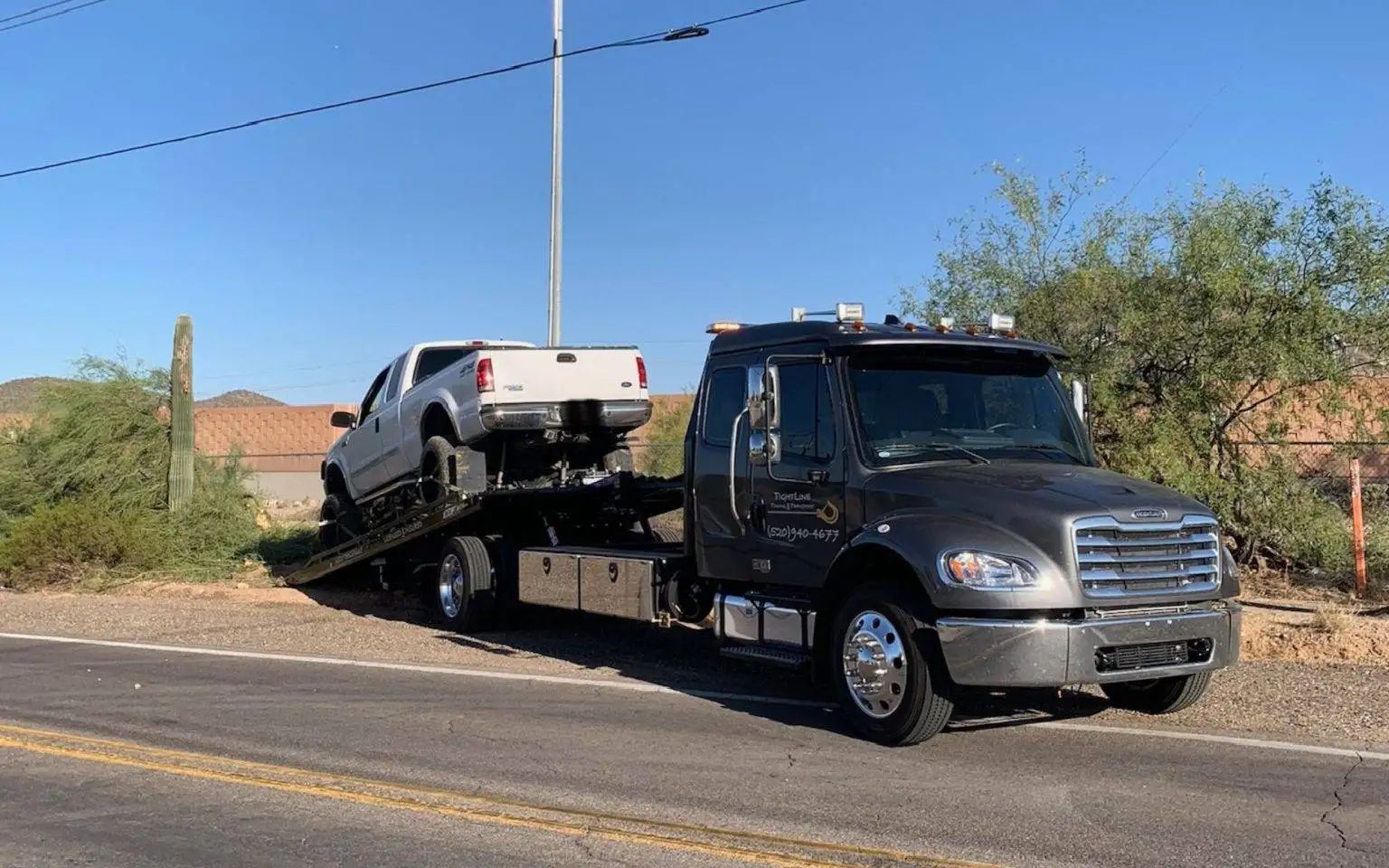 A tow truck carrying a white pickup truck on a sunny roadside.
