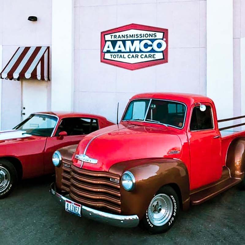 Two classic cars parked outside an AAMCO Total Car Care sign, including a red vintage pickup in front.