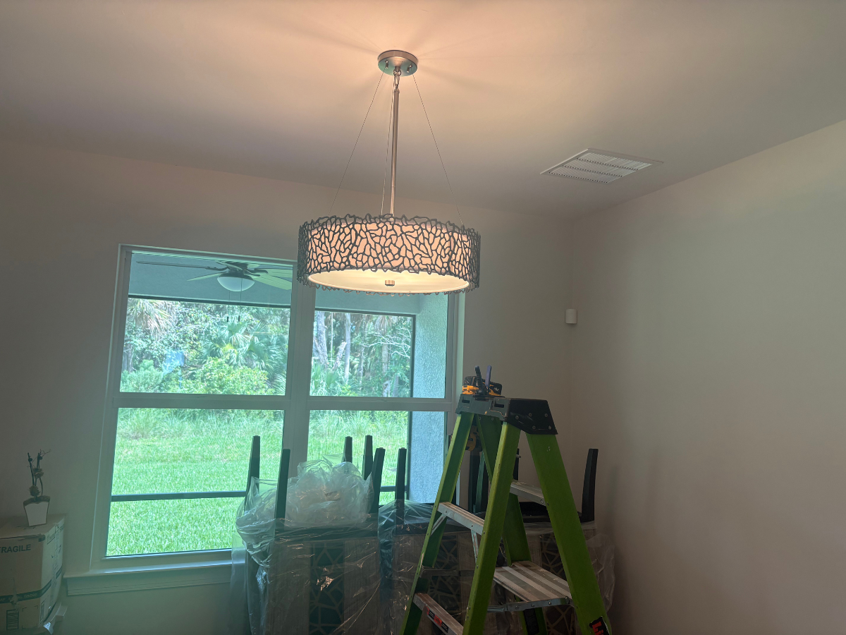 A decorative drum chandelier hangs from a light-colored ceiling above a green ladder in an unfurnished room.