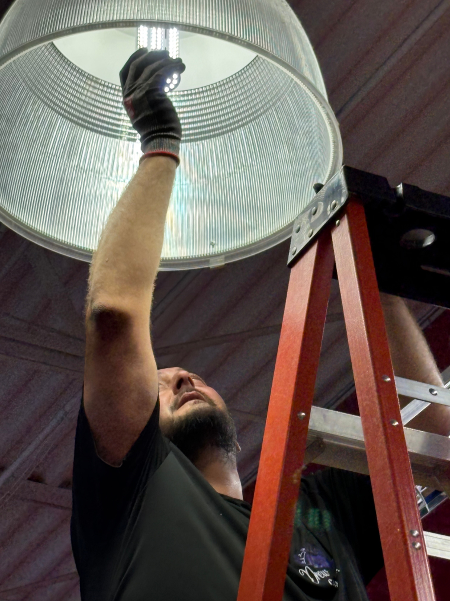 A person wearing work gloves on a red step ladder, reaching up to replace a light bulb in a large industrial dome light.