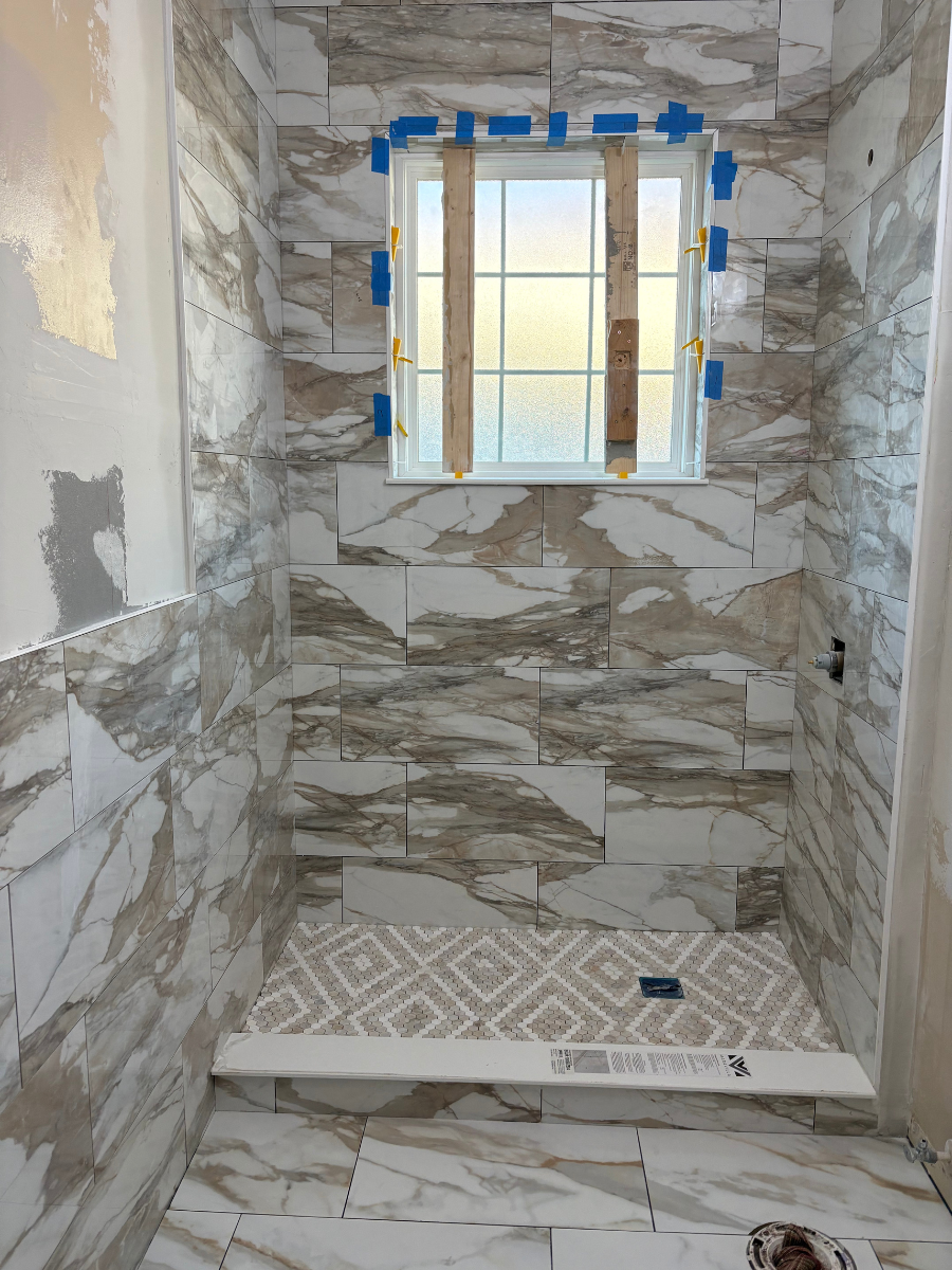 A partially tiled shower featuring marbled wall tiles, a patterned floor, and a window with blue painter's tape on the frame.