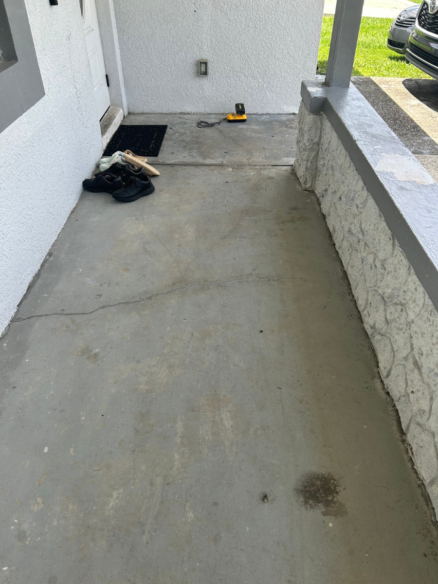 A concrete front porch with a single hairline crack, a black doormat with shoes, and a yellow power tool near the door.