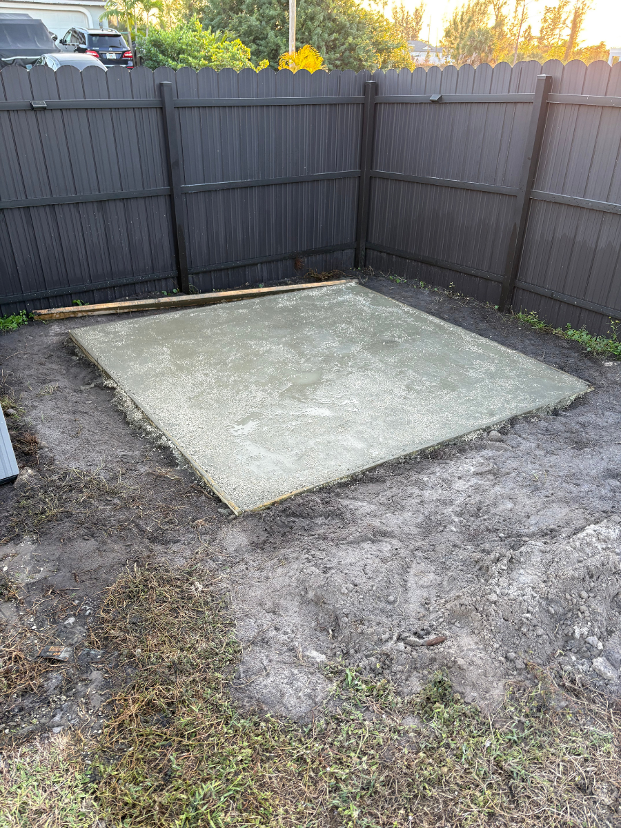 A newly poured, square concrete slab sits in a backyard corner enclosed by a dark wood fence.