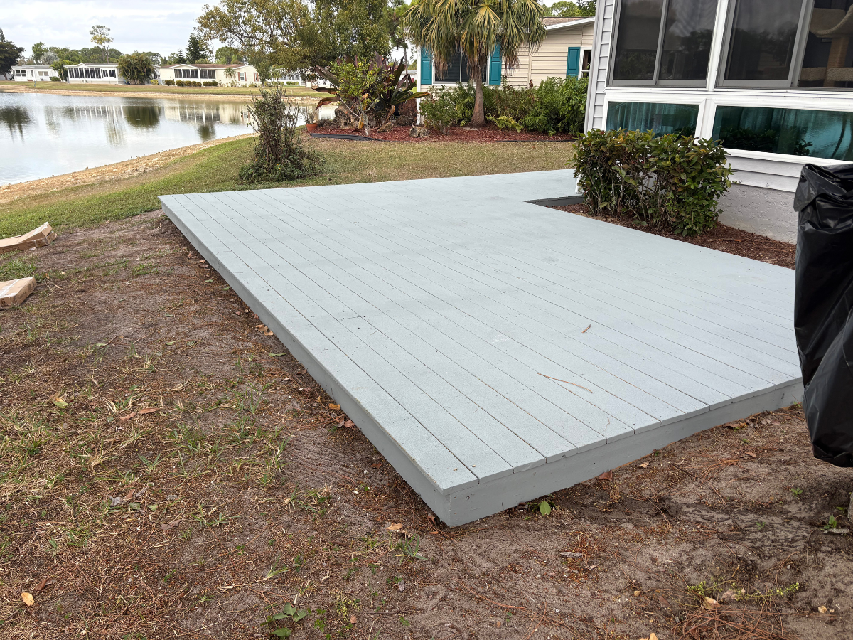 A light gray wooden deck sits in a backyard next to a house, with a pond visible in the background.