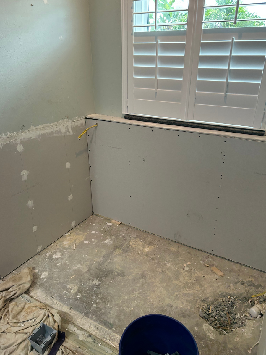 A corner of a room under renovation featuring new grey drywall panels, a window with white shutters, and a concrete floor.