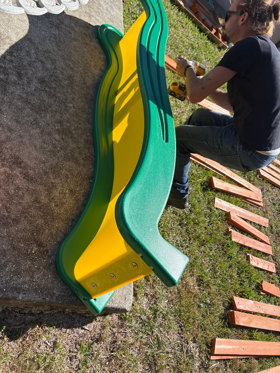 A person using a drill to attach wooden boards to a green and yellow plastic slide on a grassy lawn.