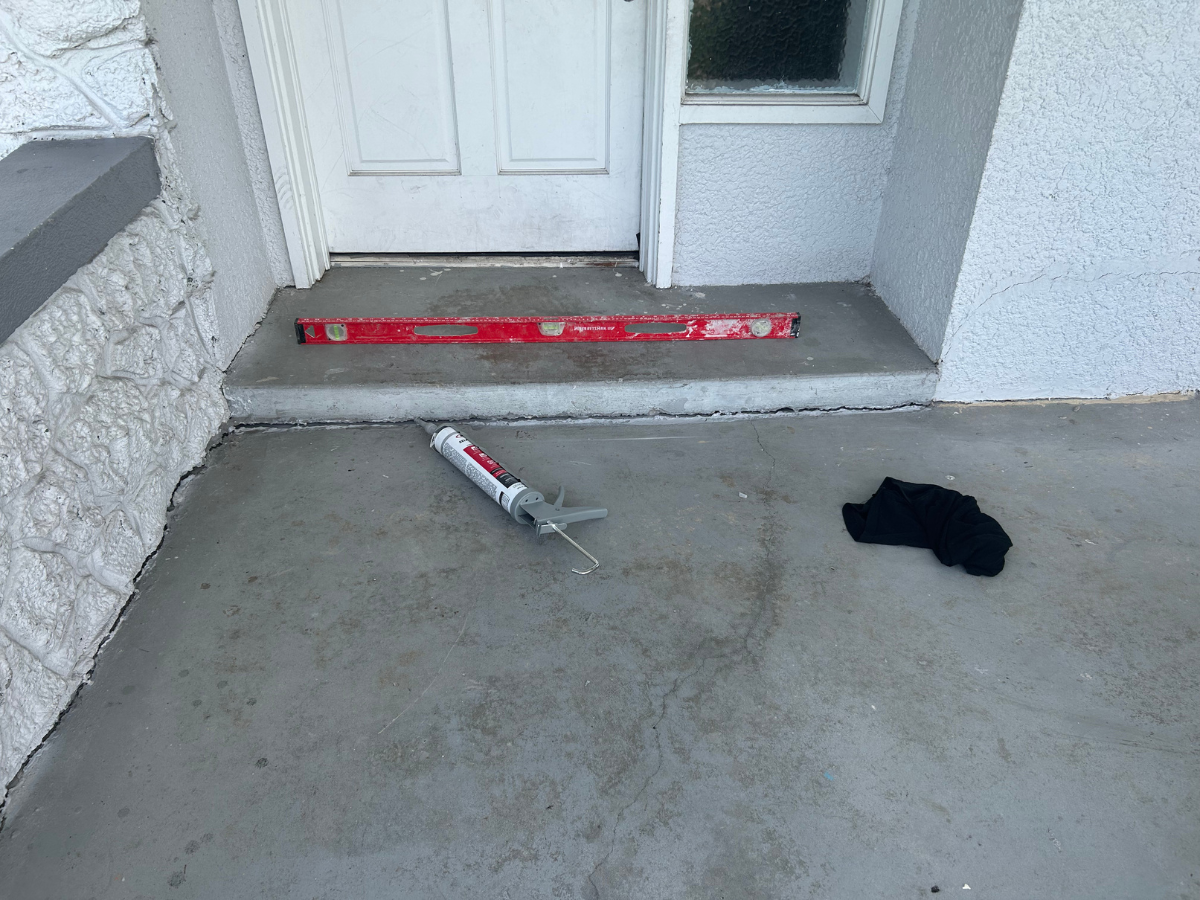 A level, a caulk gun, and a cloth rest on a concrete porch in front of an entrance door.