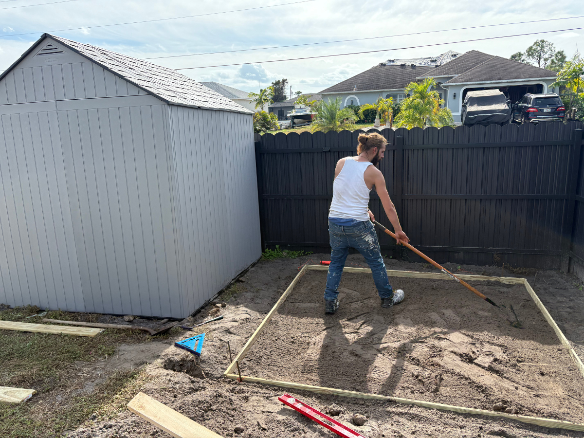 A person uses a rake to level dirt inside a wooden frame beside a shed in a backyard with a dark fence.