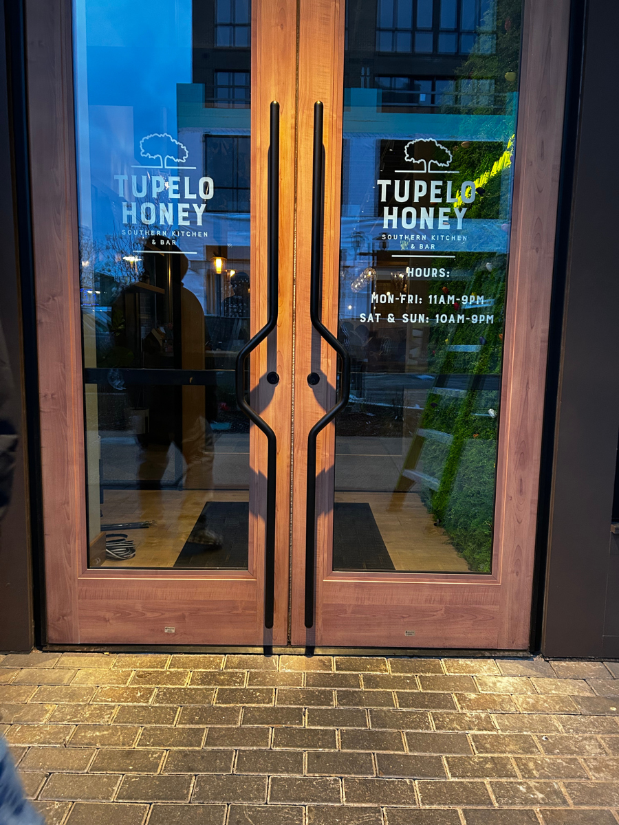 Double wooden doors to a Tupelo Honey restaurant entrance at night, featuring black vertical handles and glass windows.