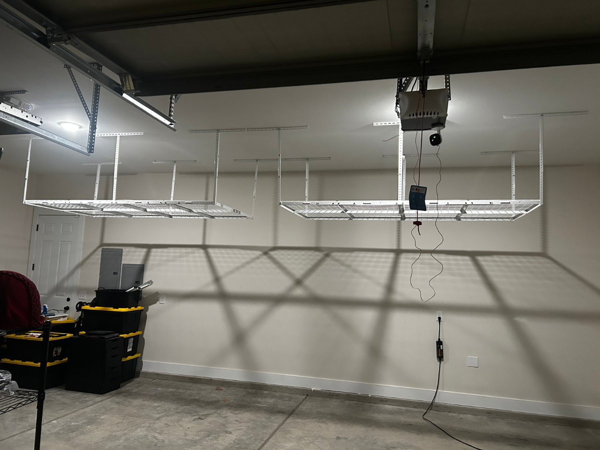 Two white metal storage racks mounted to a garage ceiling above black storage bins and a concrete floor.