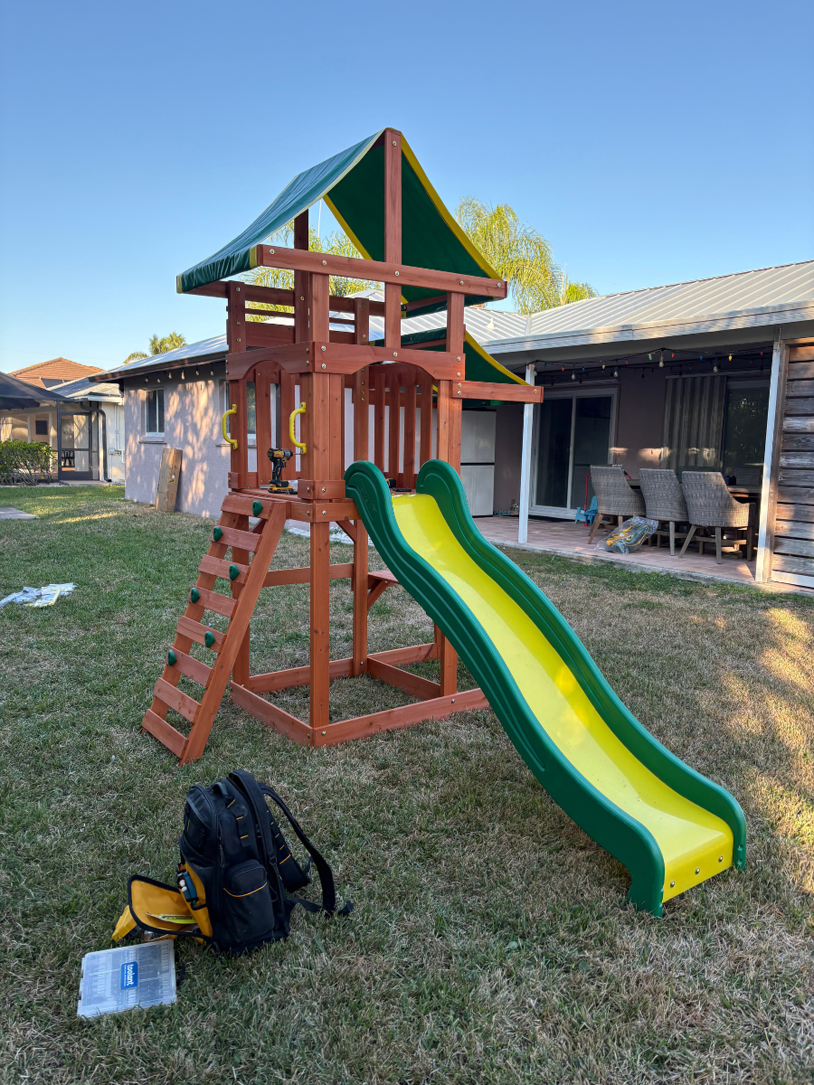 A wooden children's play set with a green roof, a yellow slide, and a climbing wall in a backyard with a backpack nearby.