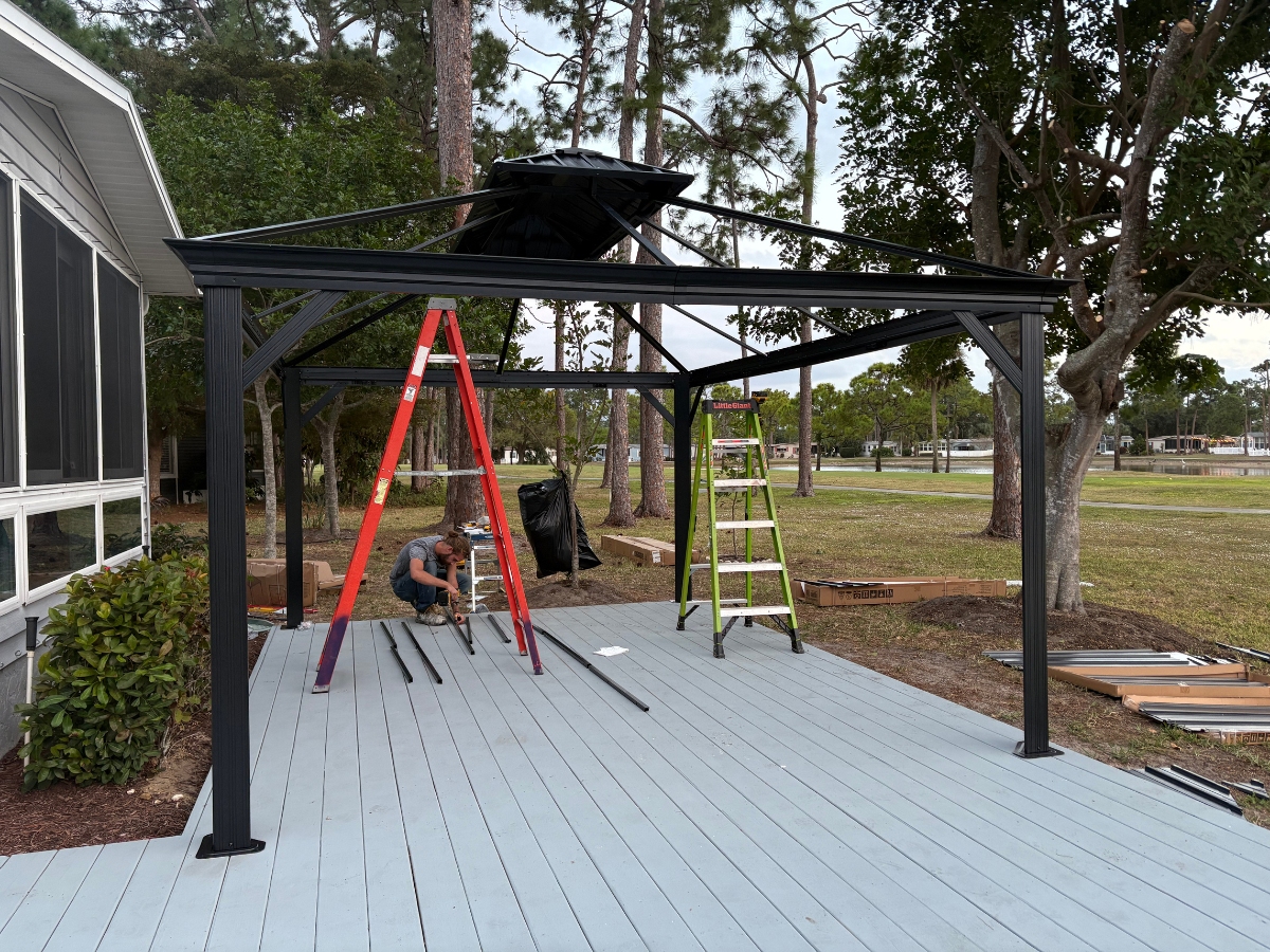 A person building a black metal gazebo frame on a light gray deck near a house, with ladders and tools nearby.