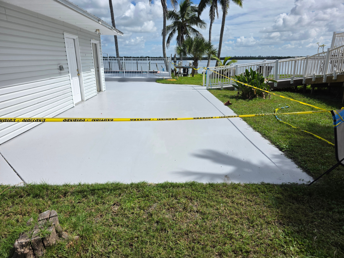 A light grey concrete patio beside a building, cordoned off by yellow caution tape, with a waterfront view in the distance.