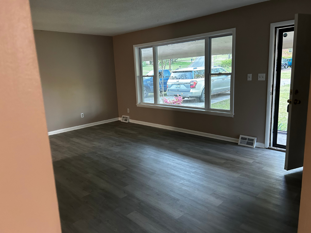 A spacious, empty living room with brown walls, dark wood-look flooring, a large window, and an open door to the outside.