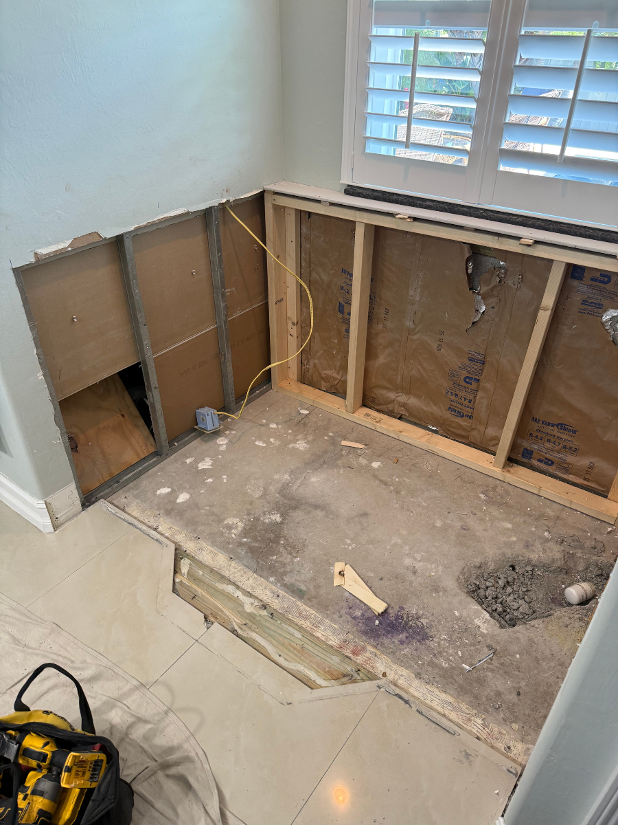 Renovation site showing an open wall with exposed wooden framing and electrical wiring above a damaged subfloor section.