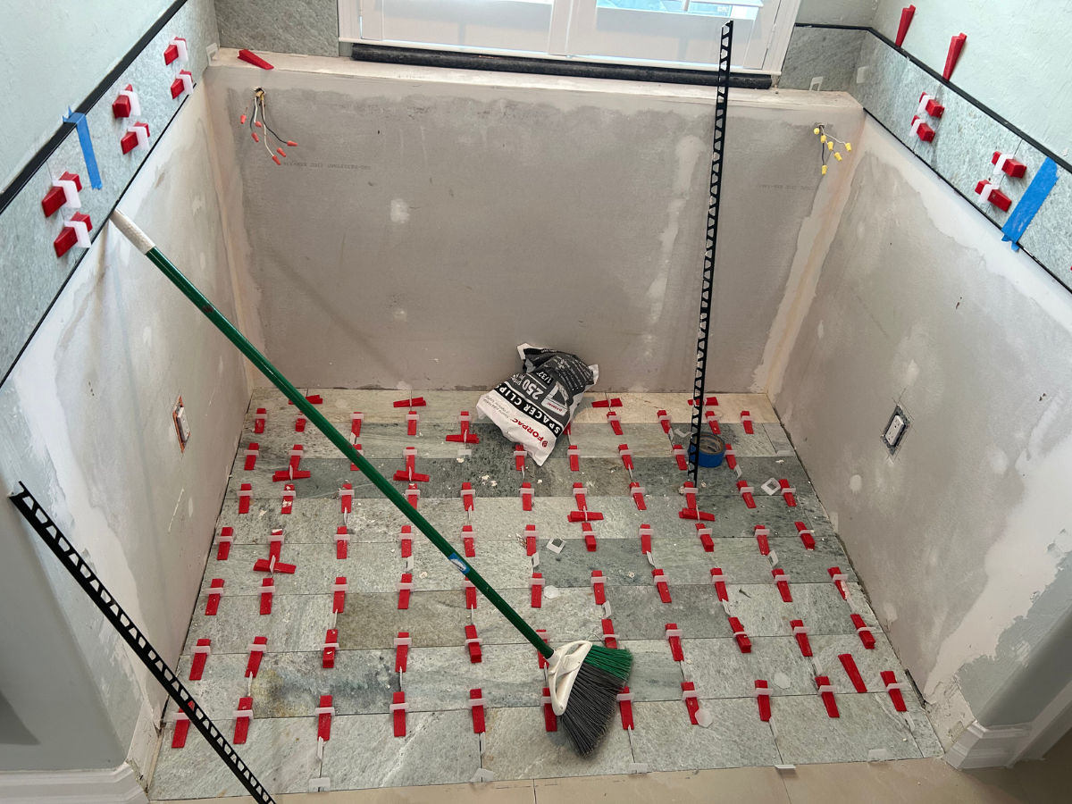 A room under construction with gray stone floor tiles held by red plastic leveling clips, wall patches, and a broom.