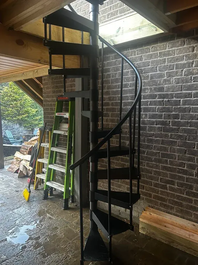 A black metal spiral staircase stands on a patio floor next to a green step ladder and a brick wall.