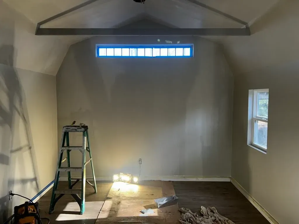 A room under renovation with beige walls, a high rectangular window, a stepladder, and flooring materials on the ground.