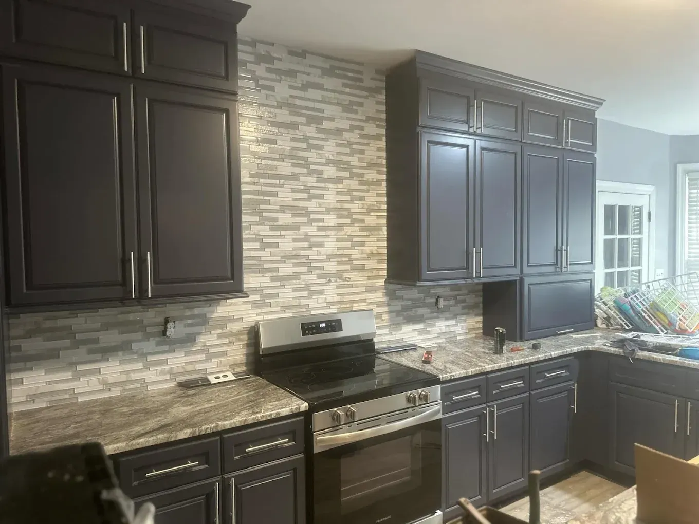 A kitchen with dark cabinets, a light-colored granite countertop, and a grey and white mosaic tile backsplash.