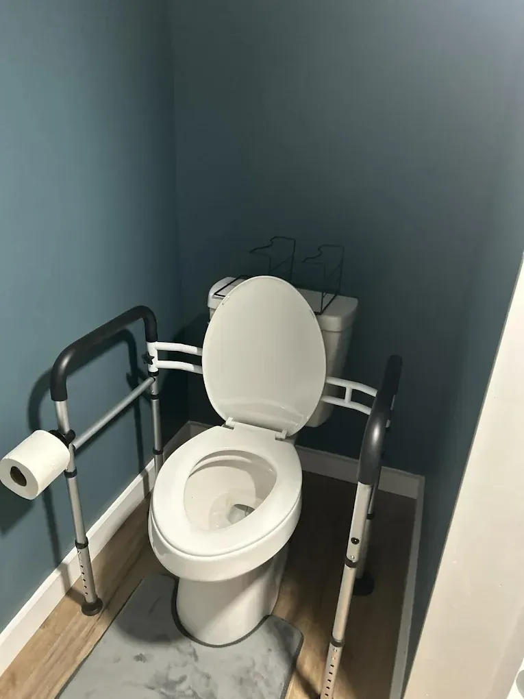 A white toilet in a small room with blue walls, equipped with a metal safety frame and toilet paper holder.