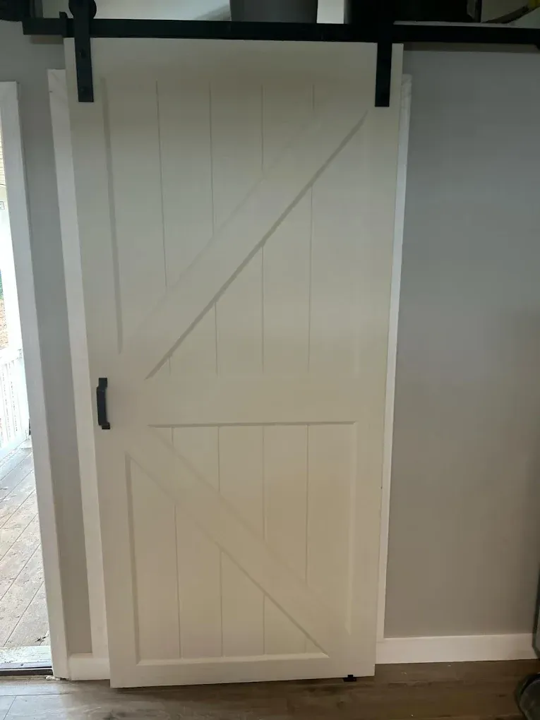 White barn door on a black sliding track; black handle. Mounted on white trim. Gray wall in the background.