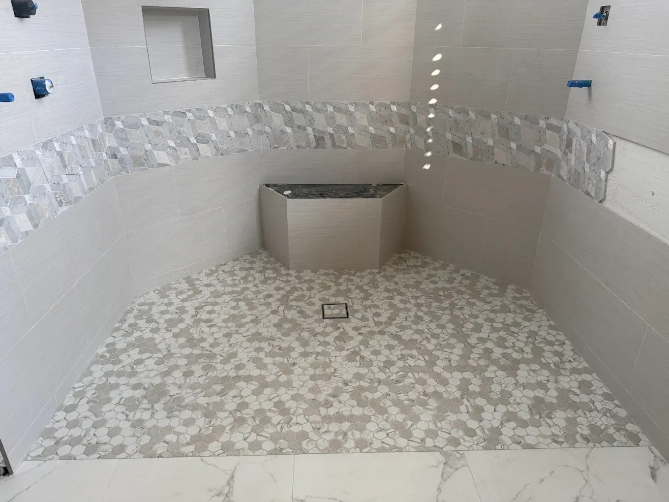 A walk-in shower under construction with white marble-look tiles, a pebble floor, a small wall niche, and plumbing fixtures.