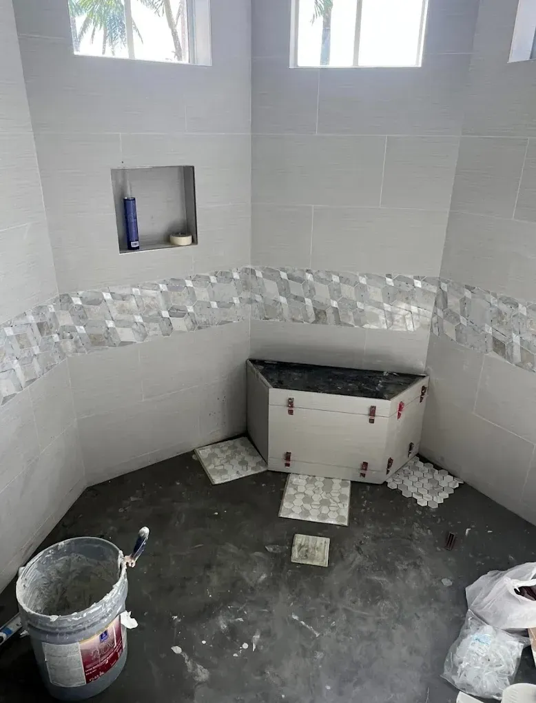 A corner shower under construction featuring grey tiles, a decorative border, a built-in niche, and floor tile samples.