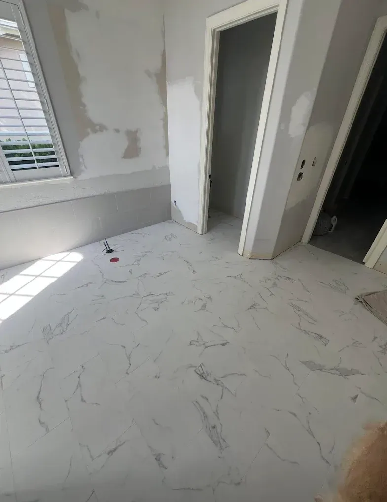 A room undergoing renovation with white marble-patterned tile flooring, unfinished drywall, and two open doorways.