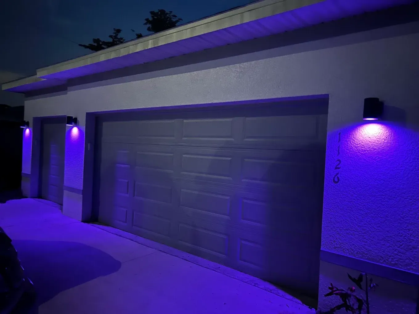 A residential garage exterior at night, illuminated by three wall-mounted sconce lights casting bright blue pools of light.