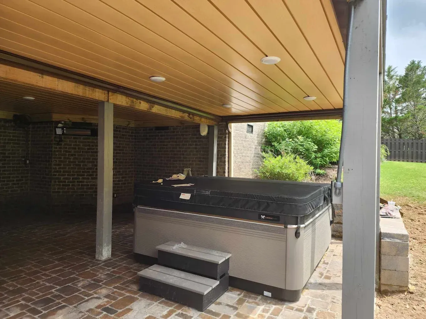 A hot tub under a covered patio with brick flooring, steps, and surrounding greenery.