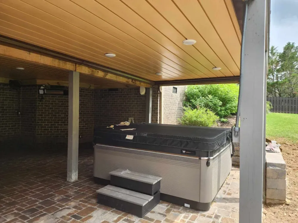 A hot tub under a covered patio with brick flooring, steps, and surrounding greenery.