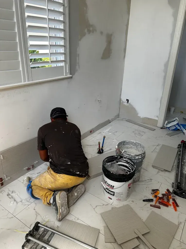 A person kneels on a white-tiled floor, installing tiles in a room with unfinished walls and buckets of supplies nearby.