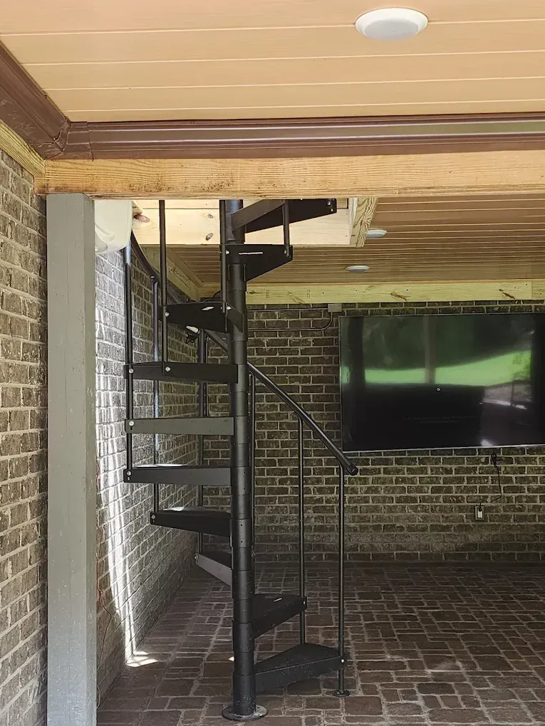 A black metal spiral staircase leads to an upper level in a covered patio with brick walls and a wooden ceiling.