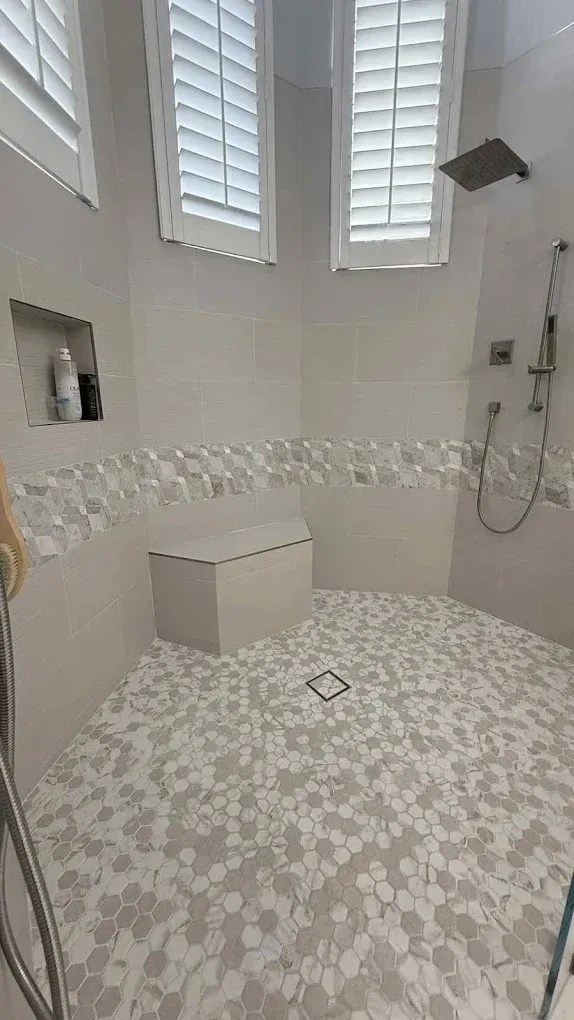 A modern walk-in shower with light grey walls, white pebble floor tiles, a built-in corner bench, and two shuttered windows.