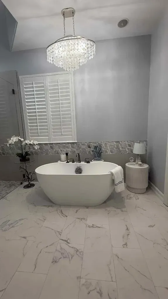A modern bathroom featuring a white freestanding tub, crystal chandelier, marble flooring, and white plantation shutters.