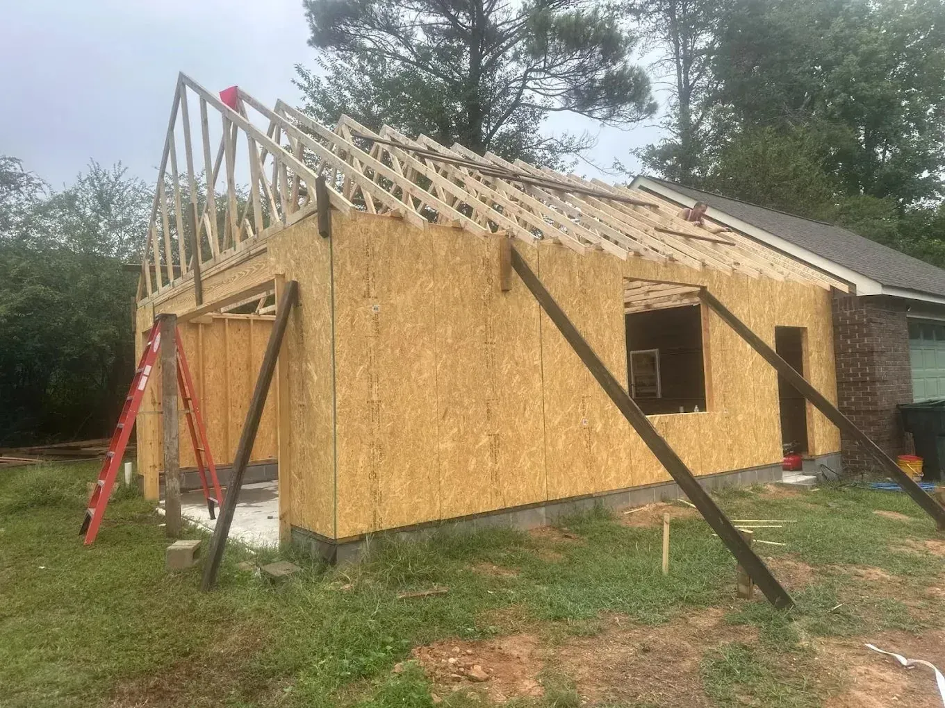 A wood-framed residential addition under construction with exposed roof trusses, OSB wall sheathing, and temporary bracing.
