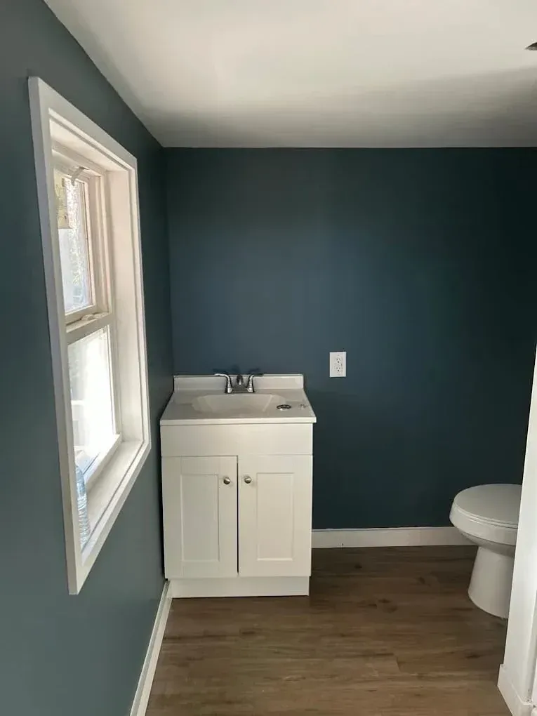 Bathroom with white sink cabinet, toilet, window, and dark teal walls.