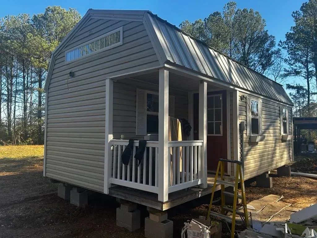 A beige, gambrel-roofed tiny home on cinder block foundations with a front porch and steps in a wooded, outdoor setting.