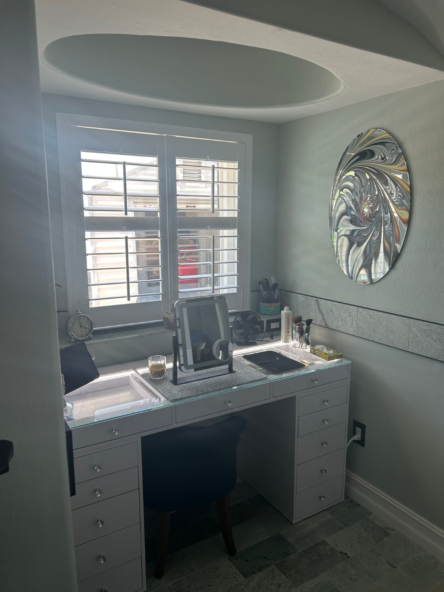 A white vanity table with drawers, a mirror, and a stool, set beneath a shuttered window in a light green room.